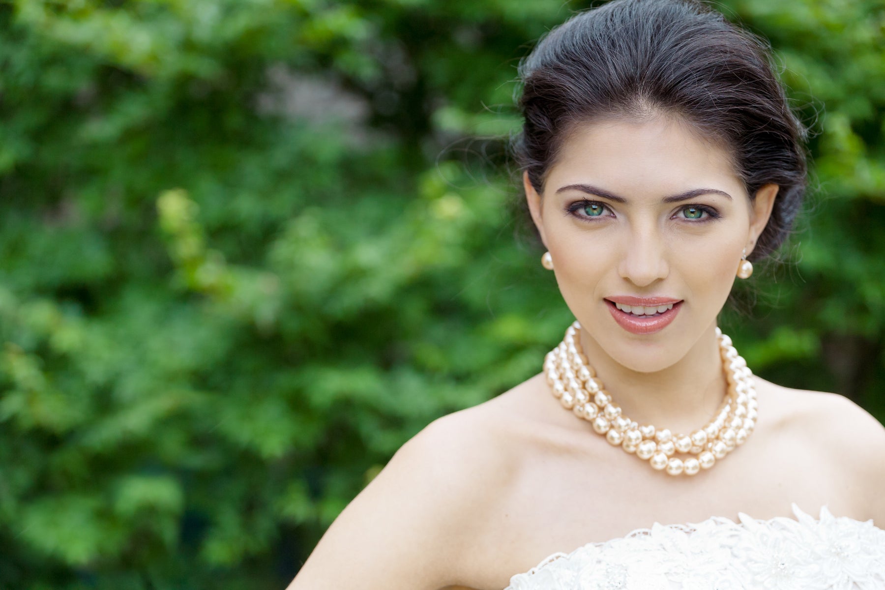 A Bride in front of green trees. The Bride is wearing blush colored pearls from Anna Bellagio.