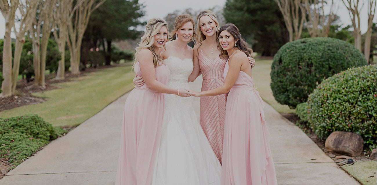 A Bride and her bridal party on a long driveway with lush green trees and bushes on both sides.